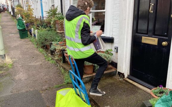 A student wearing a high visibility jacket is folding up a copy of the Exmouth Journal, ready to deliver it through the letterbox on a front door.