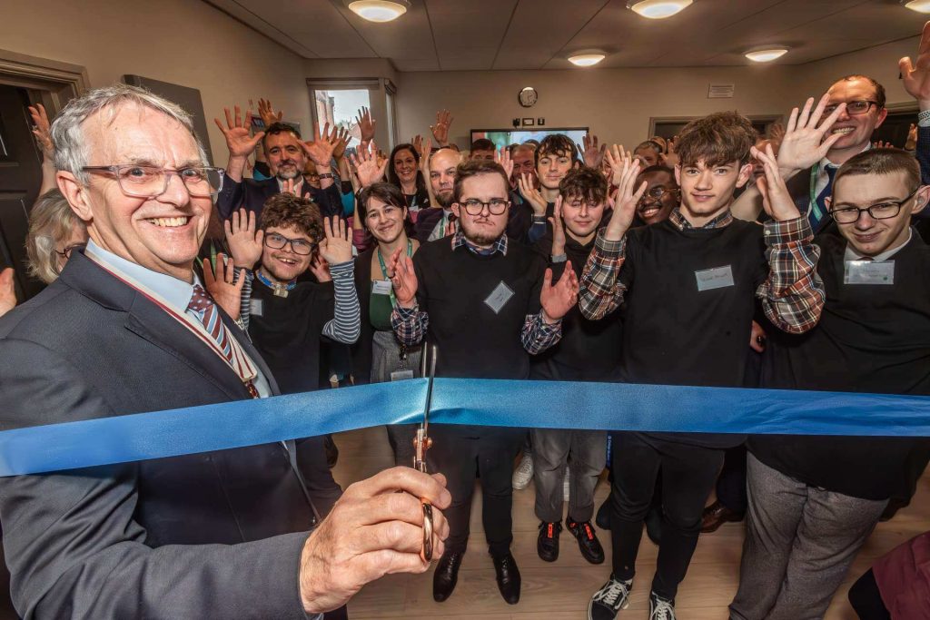 A man in the foreground on the left smiles at the camera as he cuts a blue ribbon. In the background, staff, students and guests wave their hands to in celebration.