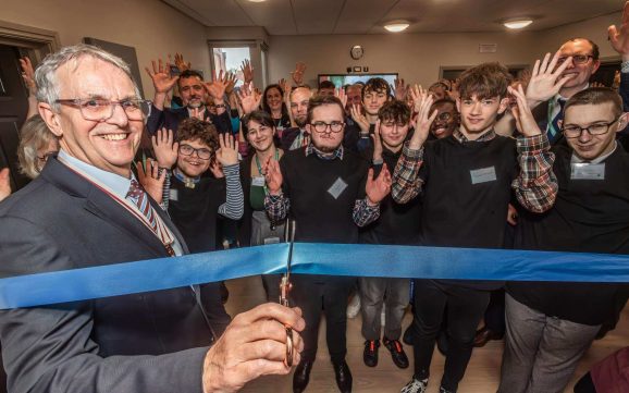 A man in the foreground on the left smiles at the camera as he cuts a blue ribbon. In the background, staff, students and guests wave their hands to in celebration.